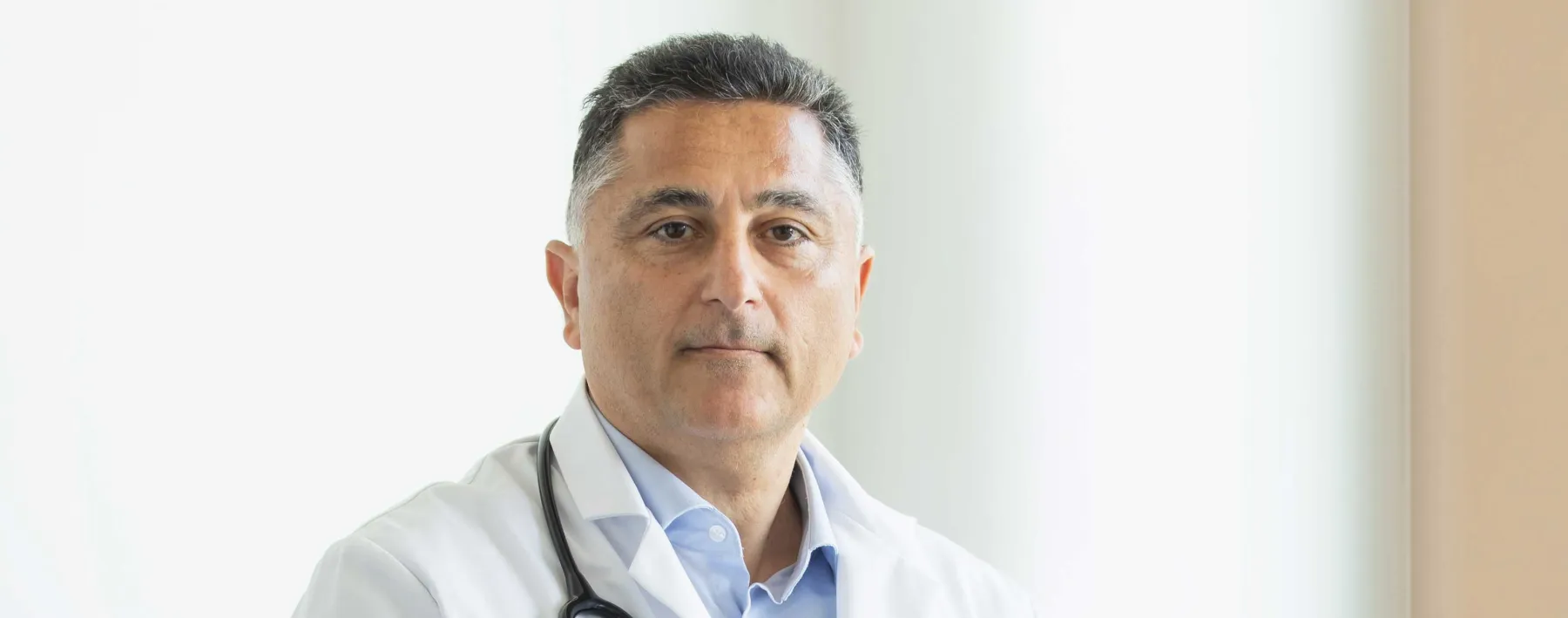 International wirkender Arzt, leicht graue Haare, mit Stethoskop um den Hals guckt in die Kamera.