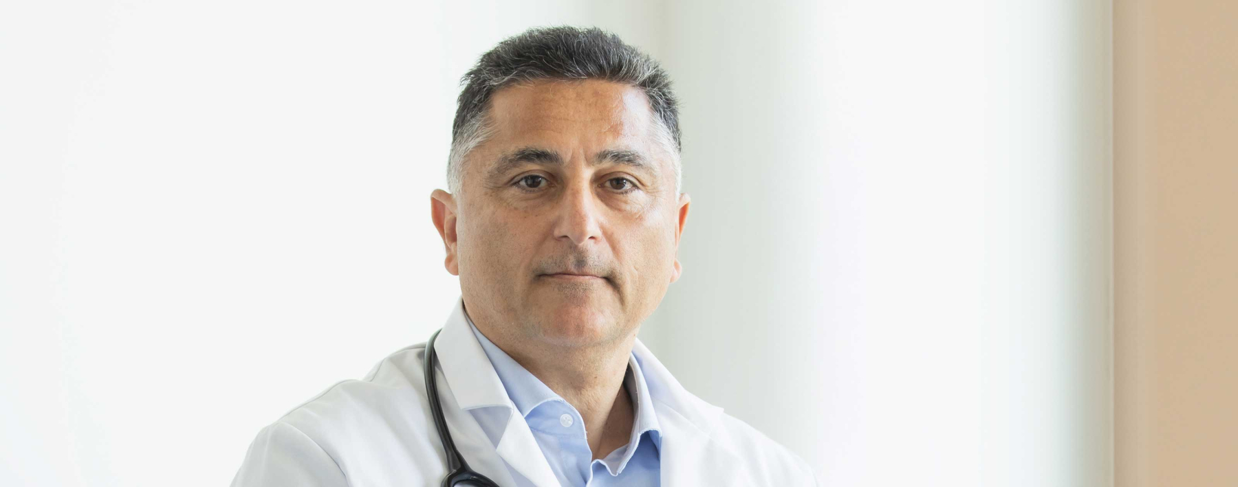 International wirkender Arzt, leicht graue Haare, mit Stethoskop um den Hals guckt in die Kamera.